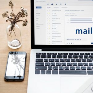 A laptop with an email inbox is displayed on the screen. The computer is on a desk, with a mobile phone and a small bouquet in a vase. A Virtual Assistant can provide admin support to small businesses.